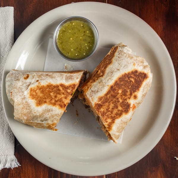A large, grilled burrito cut in half, served with a side of green salsa on a white plate.