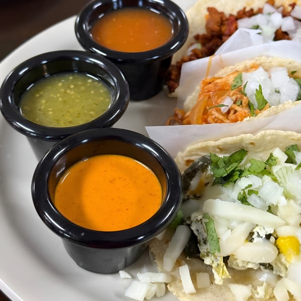 Three Tacos Godoy tacos with various fillings, topped with onions and cilantro, alongside three dipping sauces.