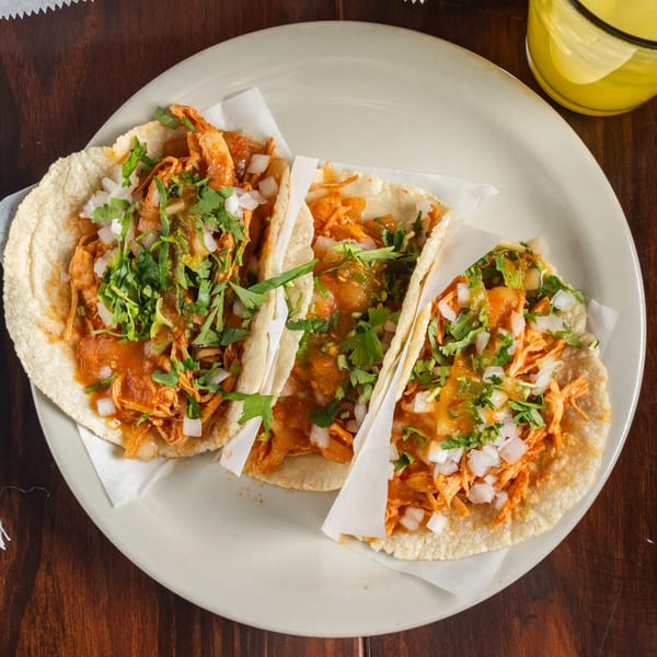 Three chicken tacos from Tacos Godoy, topped with onions, cilantro, and salsa on a white plate.