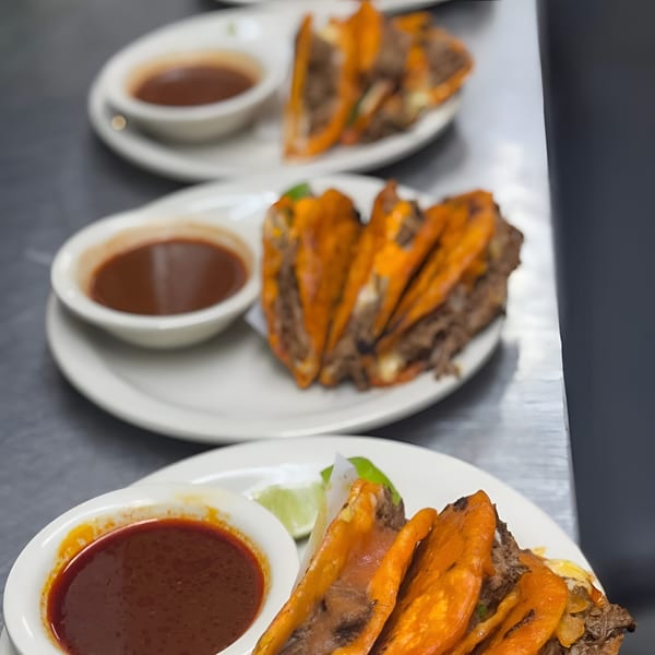 Three plates of Tacos Godoy birria tacos with dipping sauce and lime wedges on a metal counter.