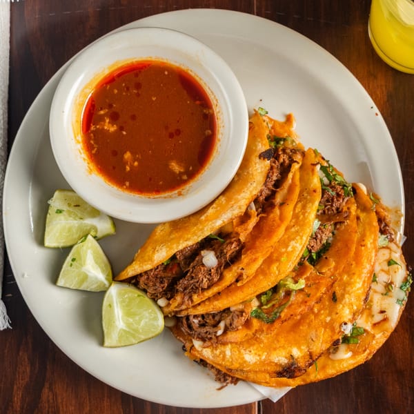 Four Tacos Godoy birria tacos with lime wedges and a side of consommé on a white plate.