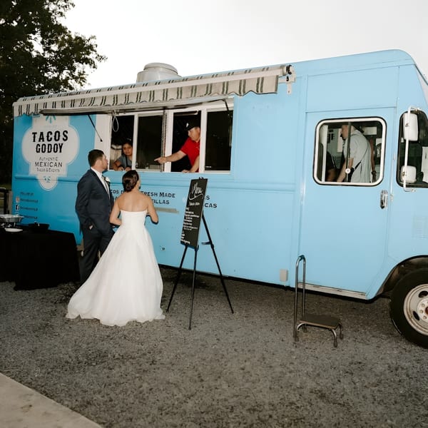 A bride and groom ordering from the Tacos Godoy food truck at an outdoor event.