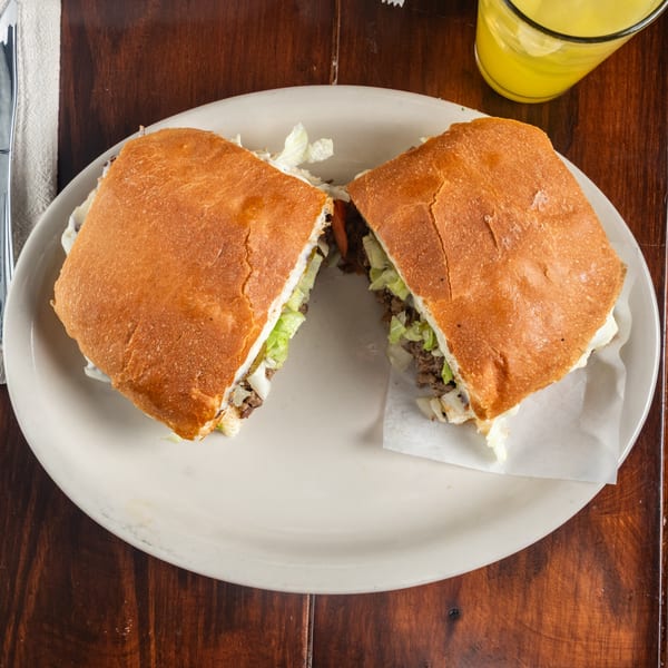 A Tacos Godoy Mexican torta, cut in half, filled with meat, lettuce, and pickles.